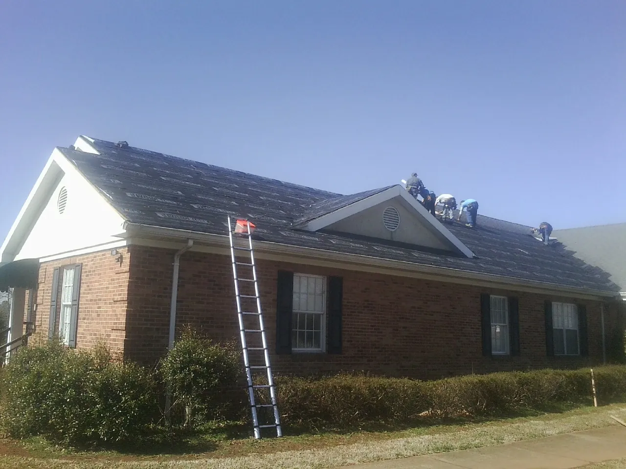 Crew tearing shingles off a brick ranch roof with a ladder set — Metal Roof Repair work by our crew in Ossian