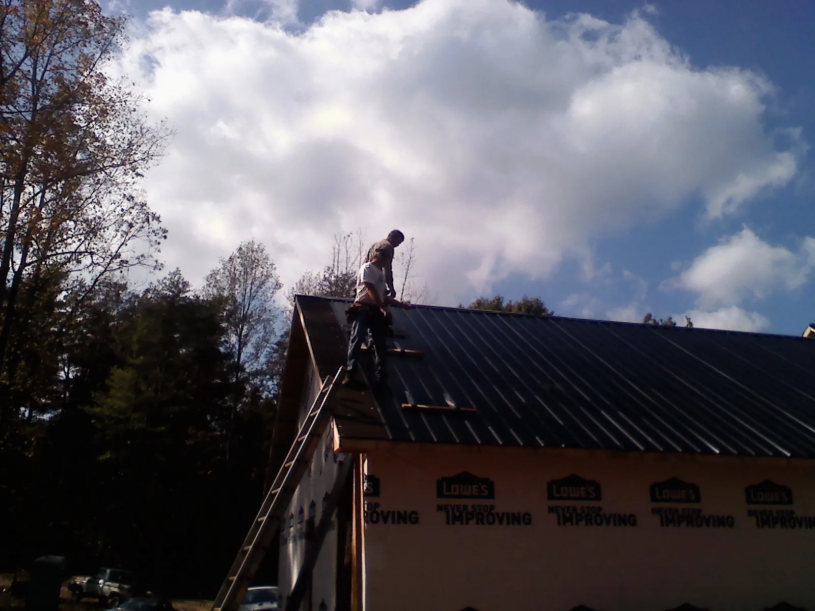 Roofer fastening dark metal panels high against a cloudy sky, from a Roof Inspection job in Ossian